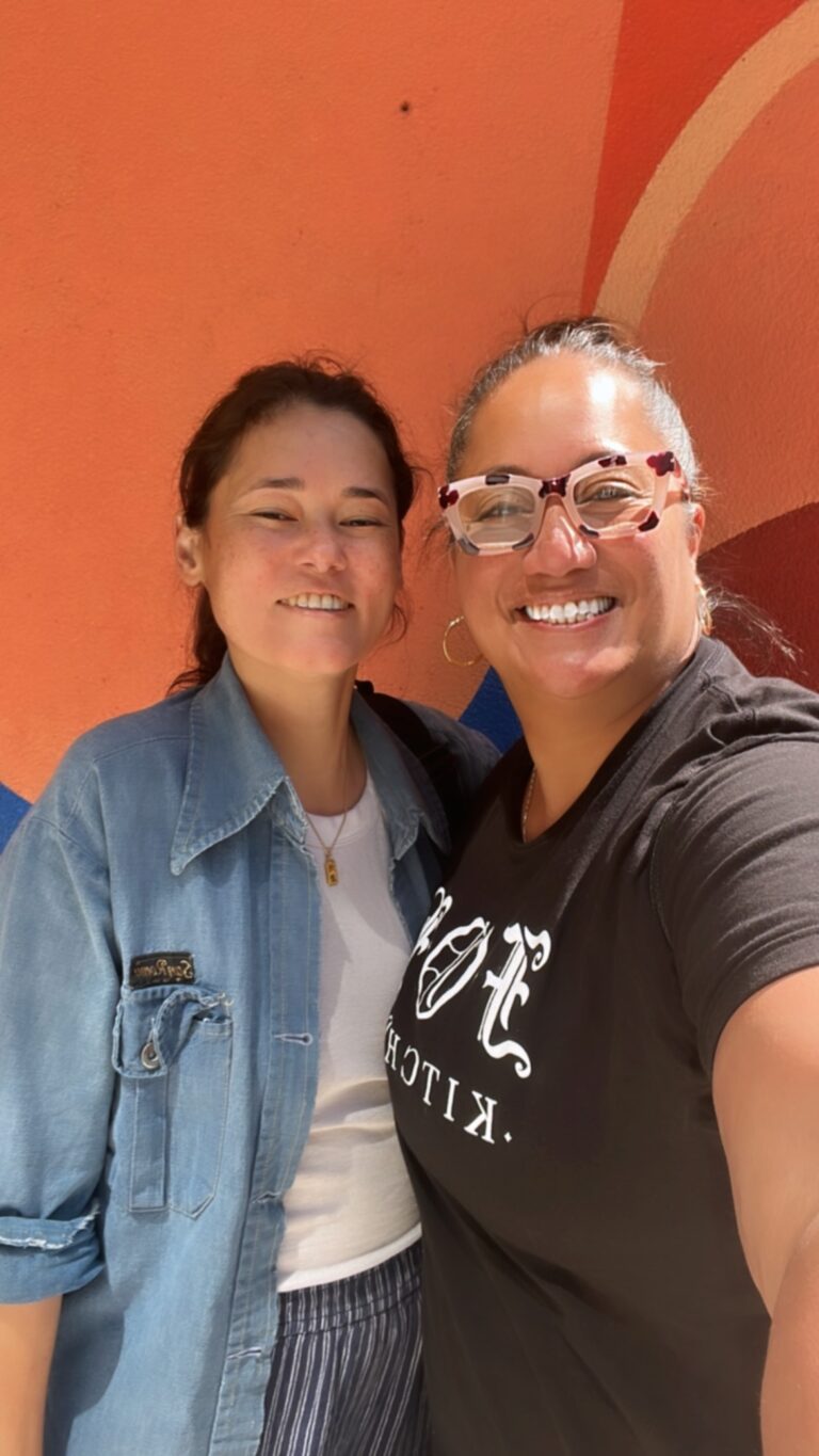 Two cis-female women standing next to each other and smiling. The woman on the left is wearing a blue denim button down shirt over a white t-shirt and pin-stripe pants. The woman on the right is wearing glasses and a black t-shirt with white text on the shirt.