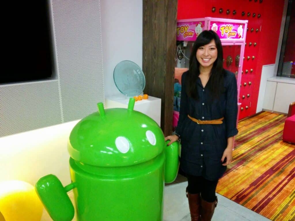 A woman with long black hair wearing a denim dress with a thin brown belt, brown boots, and black leggings holding the hand of a Google android figure.